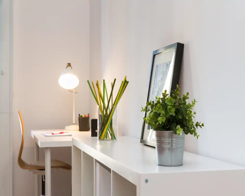 Zona giorno luminosa e minimal con una scrivania compatta e dettagli decorativi: una pianta in vaso, un quadro appoggiato e una lampada da tavolo per un angolo studio rilassante.
