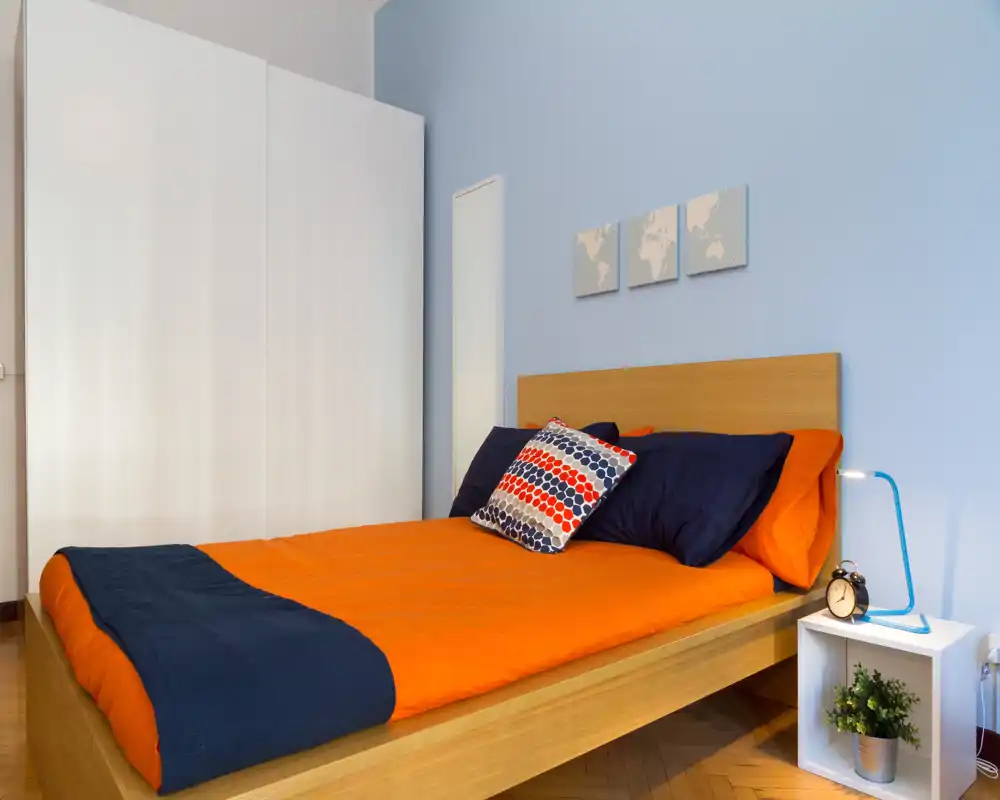 Camera da letto moderna e luminosa con letto in legno e biancheria arancione vivace con dettagli blu, armadio bianco e comodino minimal con lampada e pianta.