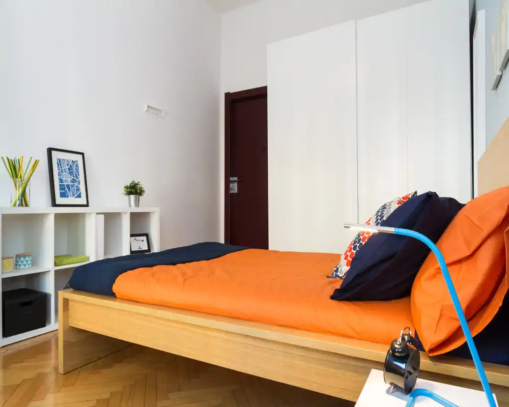 Camera da letto moderna e luminosa con letto basso in legno e biancheria arancione e blu navy, cuscini decorativi e una piccola libreria a cubi; pavimento in parquet e linee pulite creano un ambiente accogliente.
