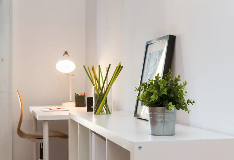 Zona giorno luminosa e minimal con una scrivania compatta e dettagli decorativi: una pianta in vaso, un quadro appoggiato e una lampada da tavolo per un angolo studio rilassante.