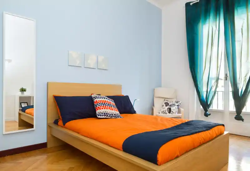 Camera da letto moderna e luminosa con letto in legno e biancheria arancione e blu navy, cuscini decorativi, specchio a figura intera e finestra con tende turchesi che lasciano entrare luce naturale.