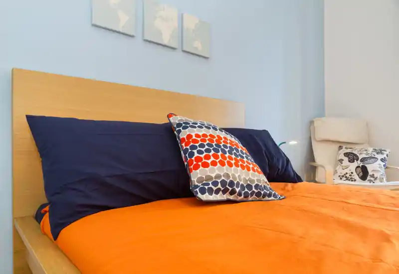 Camera da letto moderna e luminosa con testiera in legno, cuscini blu navy e una vivace coperta arancione; cuscini decorativi e una poltrona accogliente completano l'ambiente.