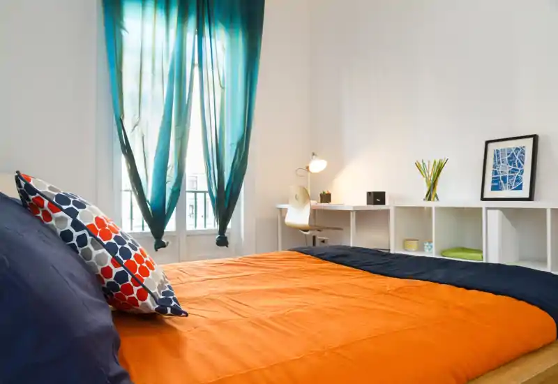 Camera luminosa e moderna con letto arancione e blu, cuscini decorativi e una scrivania vicino alla finestra — ideale per un soggiorno accogliente.