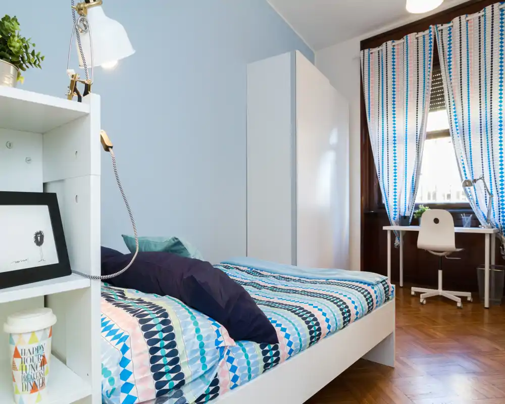 Dormitorio luminoso y bien decorado con ropa de cama estampada, una estantería junto a la cama y una zona de estudio junto a la ventana — ideal para estudiantes o inquilinos individuales.