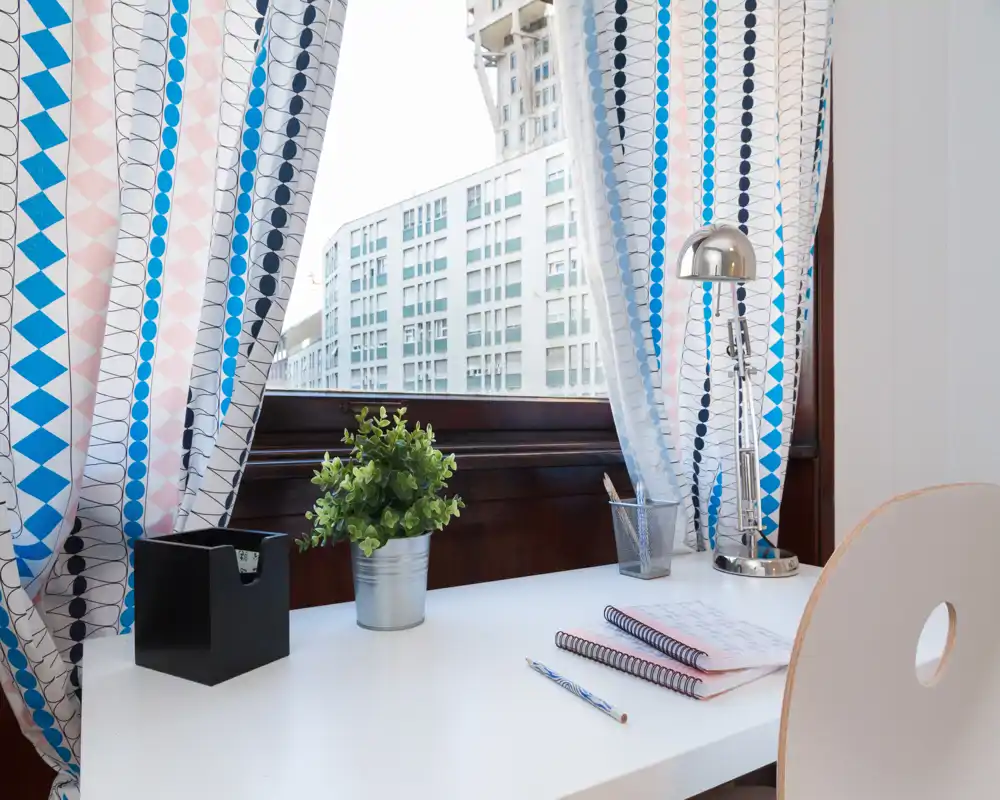 Espacio de trabajo luminoso junto a la ventana con escritorio blanco, planta decorativa, cortinas estampadas y lámpara cromada — un rincón ordenado ideal para leer o trabajar.