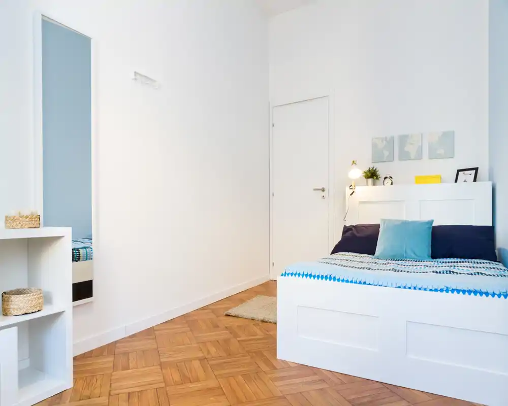 Dormitorio luminoso y minimalista con cama blanca, ropa de cama con estampado y cojines decorativos; suelo de parquet y arte de pared sencillo que crean un ambiente fresco.