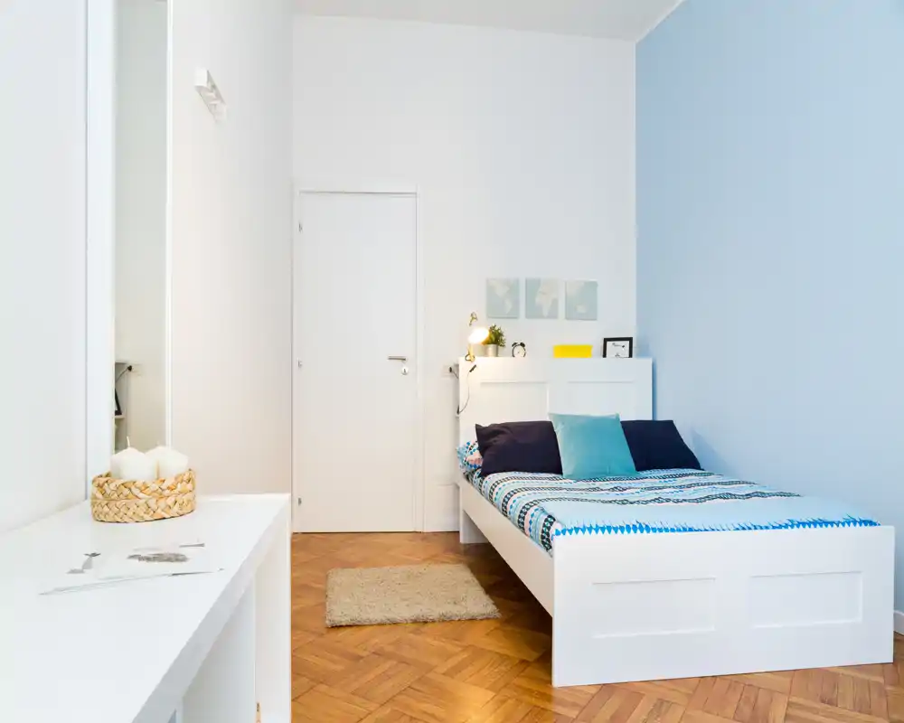 Dormitorio moderno y luminoso con estructura de cama blanca, ropa de cama con estampado y cojines decorativos; suelo de parqué limpio y una pequeña alfombra que crean un ambiente acogedor.