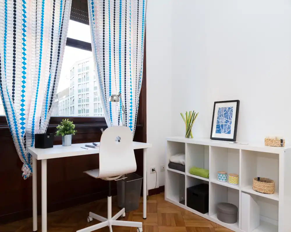 Espacio de estar luminoso y bien decorado con escritorio junto a una gran ventana, cortinas con estampado y estantería modular — ideal para teletrabajo o estudio.