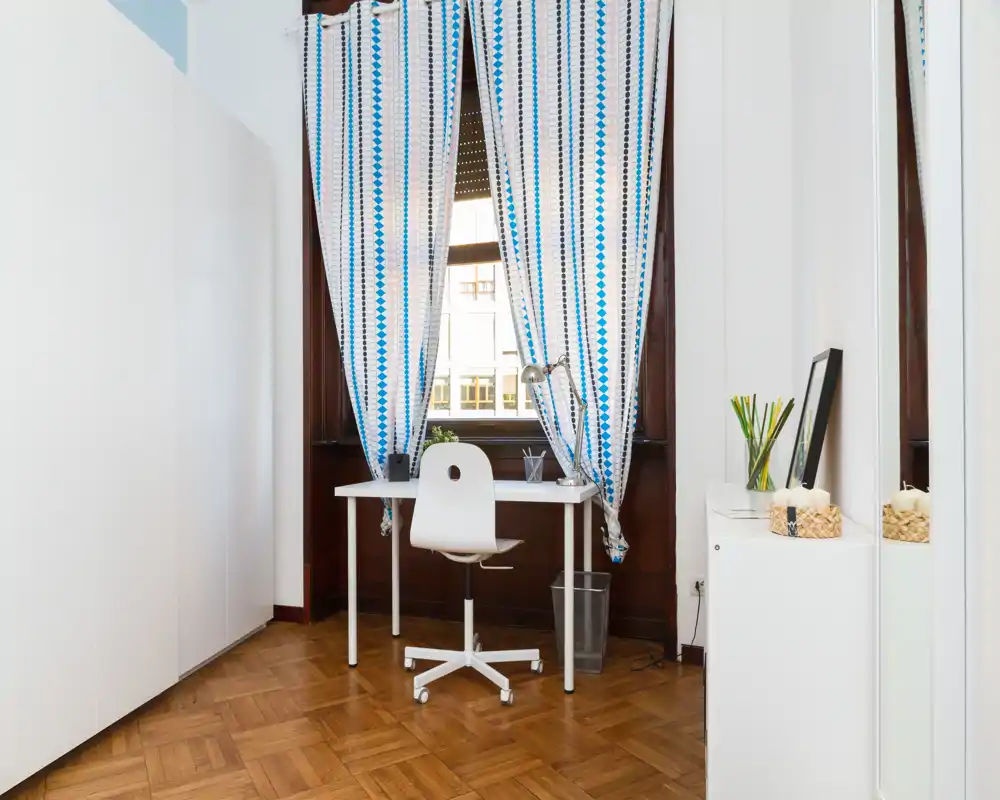 Rincón de la sala bien iluminado acondicionado como oficina en casa con escritorio blanco, silla giratoria y ventana alta con cortinas estampadas. Suelo de parquet, armario y pocos detalles decorativos crean un espacio limpio y funcional.