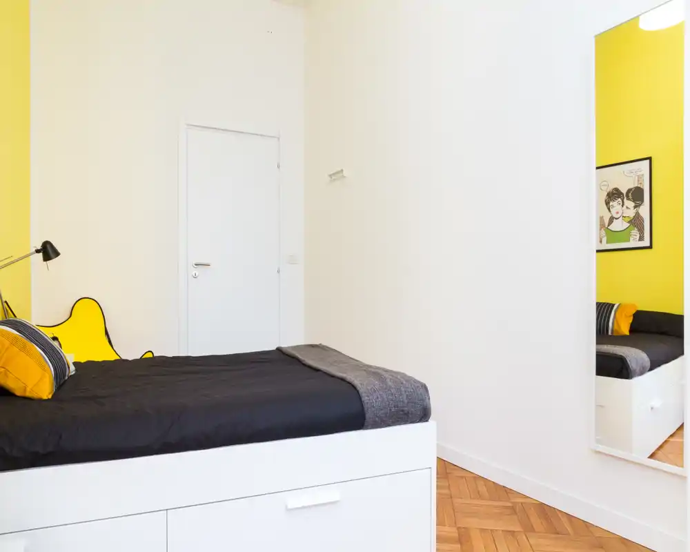 Camera da letto luminosa e moderna con letto a piattaforma bianco, biancheria nera e parete d'accento gialla. Stile minimal pulito, cuscini accoglienti e luce naturale nitida; perfetta per l'annuncio di un affitto.
