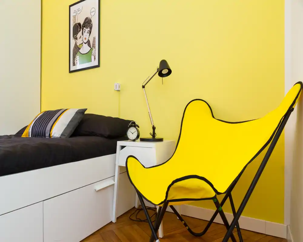 Angolo camera luminoso e moderno con letto nero, comodino e una sedia gialla d'accento su una parete gialla vivace.