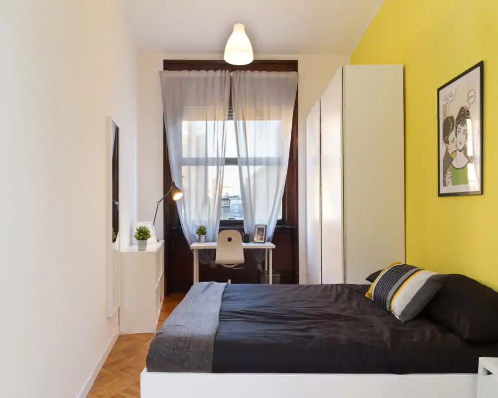 Camera da letto moderna e luminosa con letto matrimoniale in biancheria scura, una postazione di lavoro vicino alla finestra, armadio e parete accentata gialla che crea un’atmosfera accogliente.