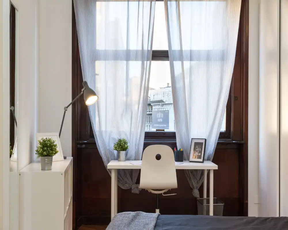 Angolo della camera luminoso e ben curato con una scrivania davanti a una grande finestra. Si vede una parte del letto in primo piano, una postazione di lavoro con sedia, lampada e piante decorative che creano un'atmosfera accogliente.