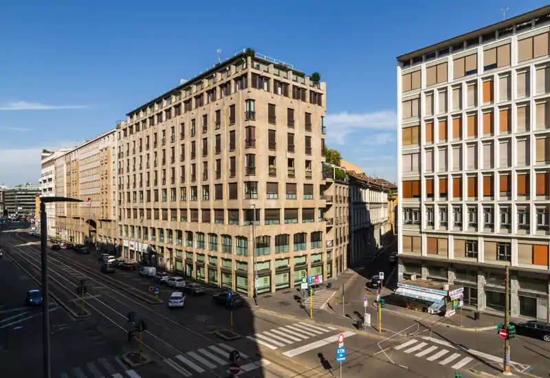 Ampia vista cittadina dall'alto con un edificio d'angolo, binari del tram e cielo terso — perfetta per evidenziare la posizione urbana e i collegamenti.