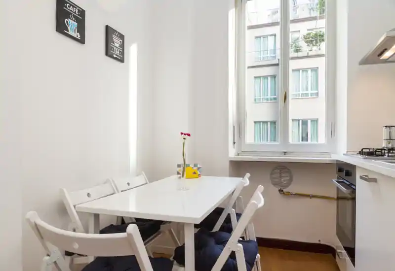 Angolo pranzo compatto e luminoso con tavolo bianco e sedie pieghevoli accanto a una grande finestra; centrotavola semplice e cuscini creano uno spazio funzionale e ordinato.