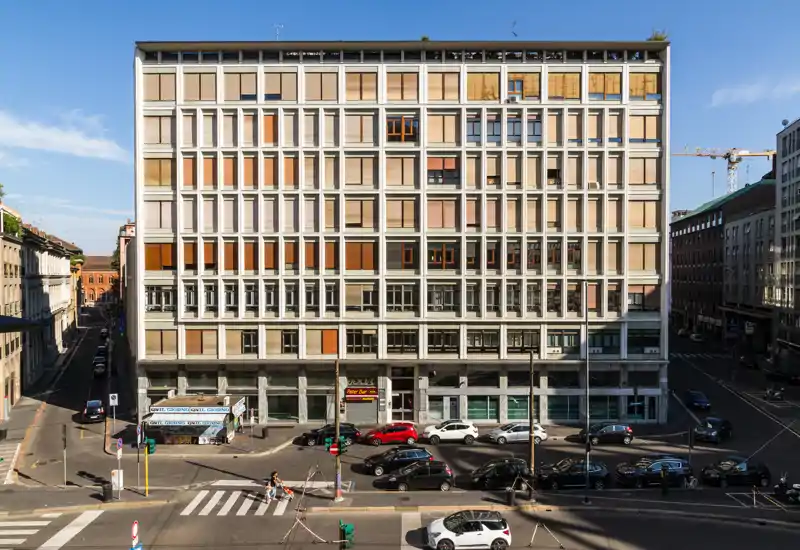 Facciata esterna su strada fotografata dall'altro lato della carreggiata, con un edificio di più piani, finestre e tapparelle e auto parcheggiate lungo la strada.