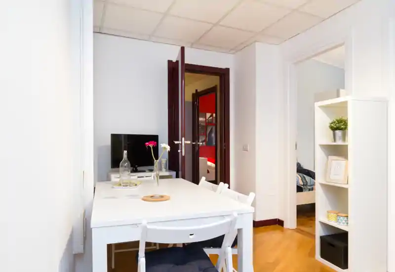 Zona giorno compatta e luminosa con tavolo bianco, sedie e una TV; la porta aperta mostra la camera adiacente. Stile moderno e dettagli decorativi che rendono l'ambiente accogliente.