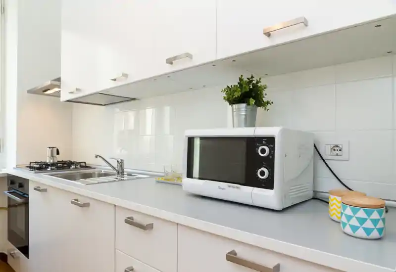 Cucinotto moderno e luminoso con mobili bianchi, lavello e piano cottura integrati, microonde sul piano e barattoli decorativi; compatto e ben organizzato.