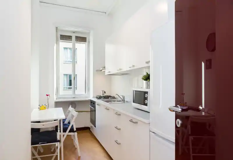 Cucina moderna compatta con mobili bianchi, piano cottura a gas, microonde e piccolo tavolo da pranzo vicino a una finestra luminosa — ideale per un appartamento in città.