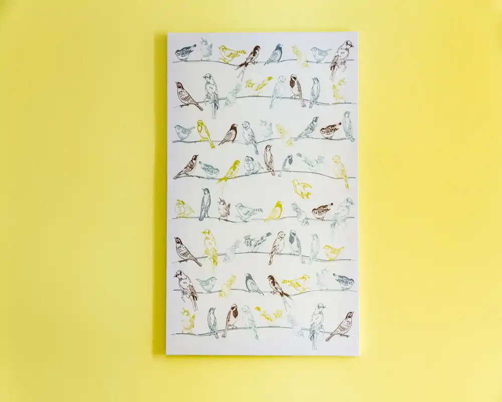 Arte de pared: un lienzo/poster vertical con múltiples pequeñas ilustraciones de pájaros colgado en una pared amarilla brillante.