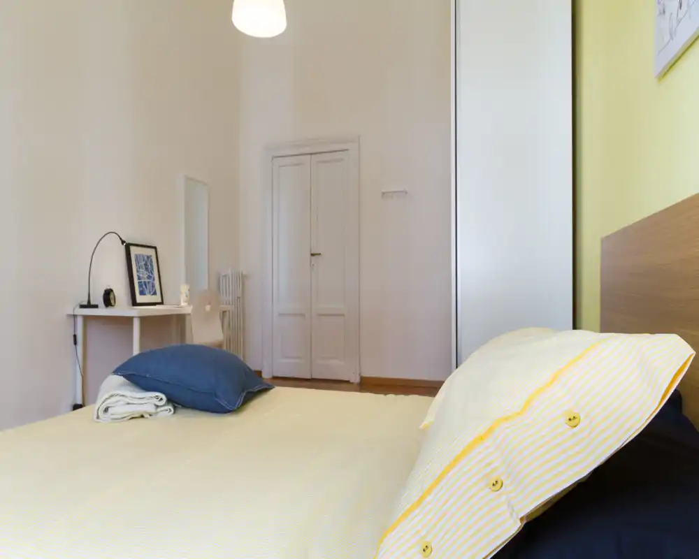 Dormitorio luminoso y ordenado con almohada de rayas amarillas y ropa de cama suave sobre una cabecera de madera baja. Se aprecia un pequeño escritorio con lámpara y un armario, un espacio para dormir limpio y acogedor.