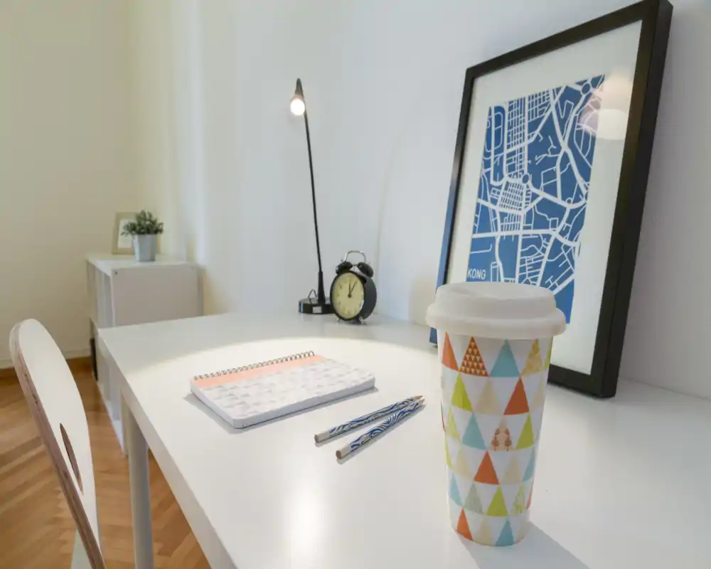 Rincón de escritorio luminoso y minimalista con vaso de viaje estampado, libreta, bolígrafos, lámpara y cuadro enmarcado: un espacio de trabajo ordenado ideal para estancias cortas.