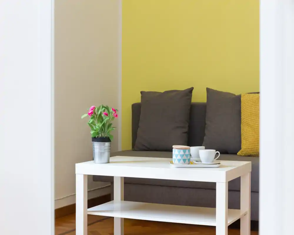 Zona giorno luminosa e moderna con divano grigio, cuscini decorativi e un tavolino bianco con tazzine e una piantina — un angolo accogliente per rilassarsi.