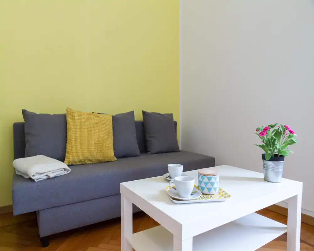 Zona giorno luminosa e moderna con divano grigio, cuscino giallo come accento e un tavolino bianco con tazze e una piccola pianta.