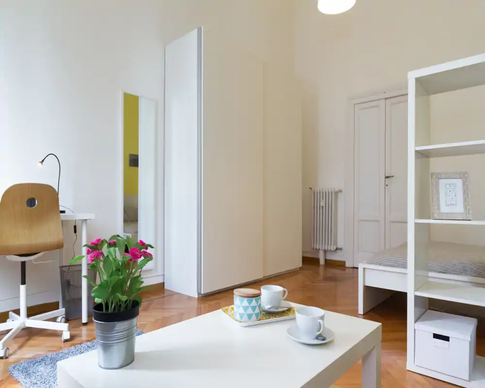 Camera da letto luminosa e minimale con letto singolo, armadio bianco e un piccolo tavolino. Pavimento in parquet, angolo scrivania e piante decorative per un’atmosfera fresca e ariosa.