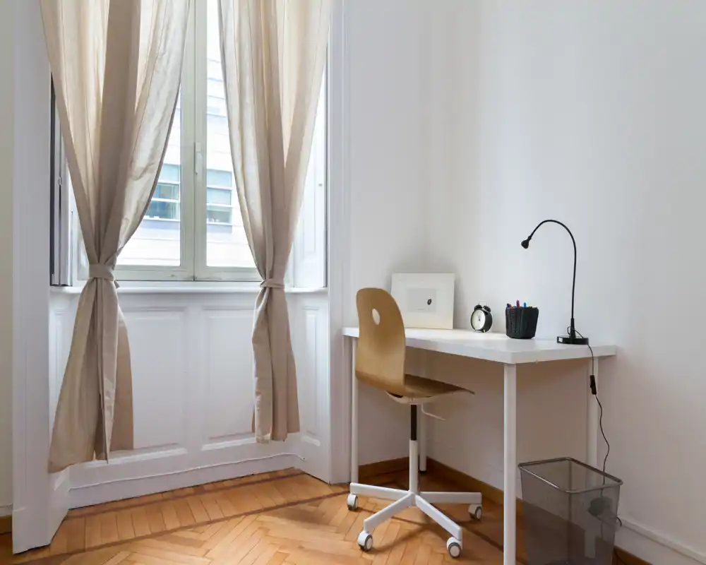 Angolo studio compatto e luminoso con scrivania bianca minimal, sedia girevole in legno, lampada da tavolo e tende beige chiare davanti a una grande finestra.