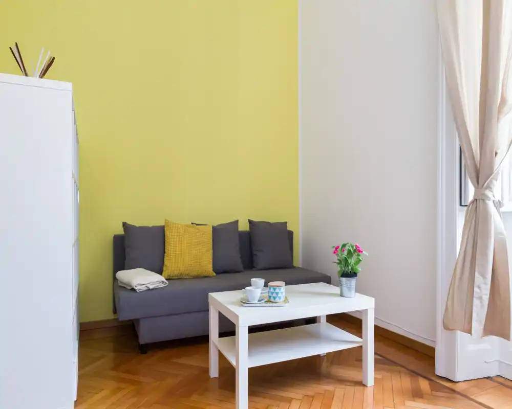 Zona giorno luminosa e moderna con un divano grigio compatto, cuscino giallo a contrasto e un tavolino bianco su pavimento in parquet caldo. La luce naturale dalla finestra e l’arredo curato creano un ambiente accogliente ideale per affitti brevi.