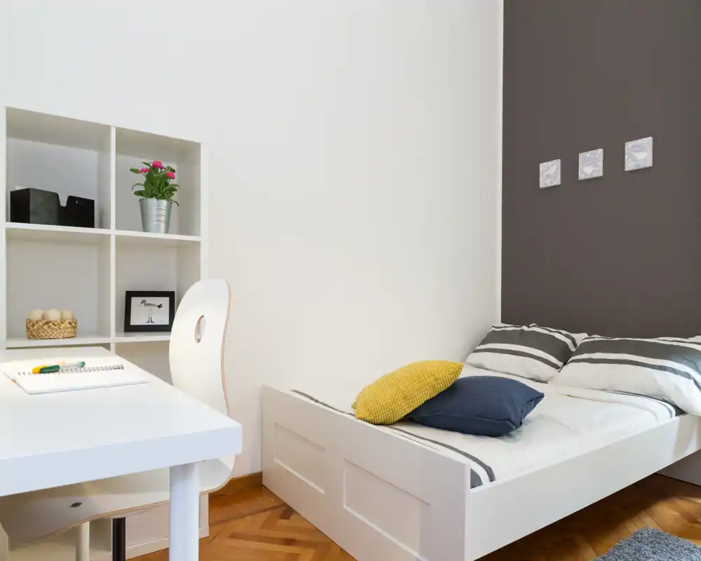 Chambre simple moderne et lumineuse avec lit à cadre blanc, linge de lit rayé et coussins d'accent; comprend un petit bureau blanc et des étagères, idéal pour étudier ou télétravailler.