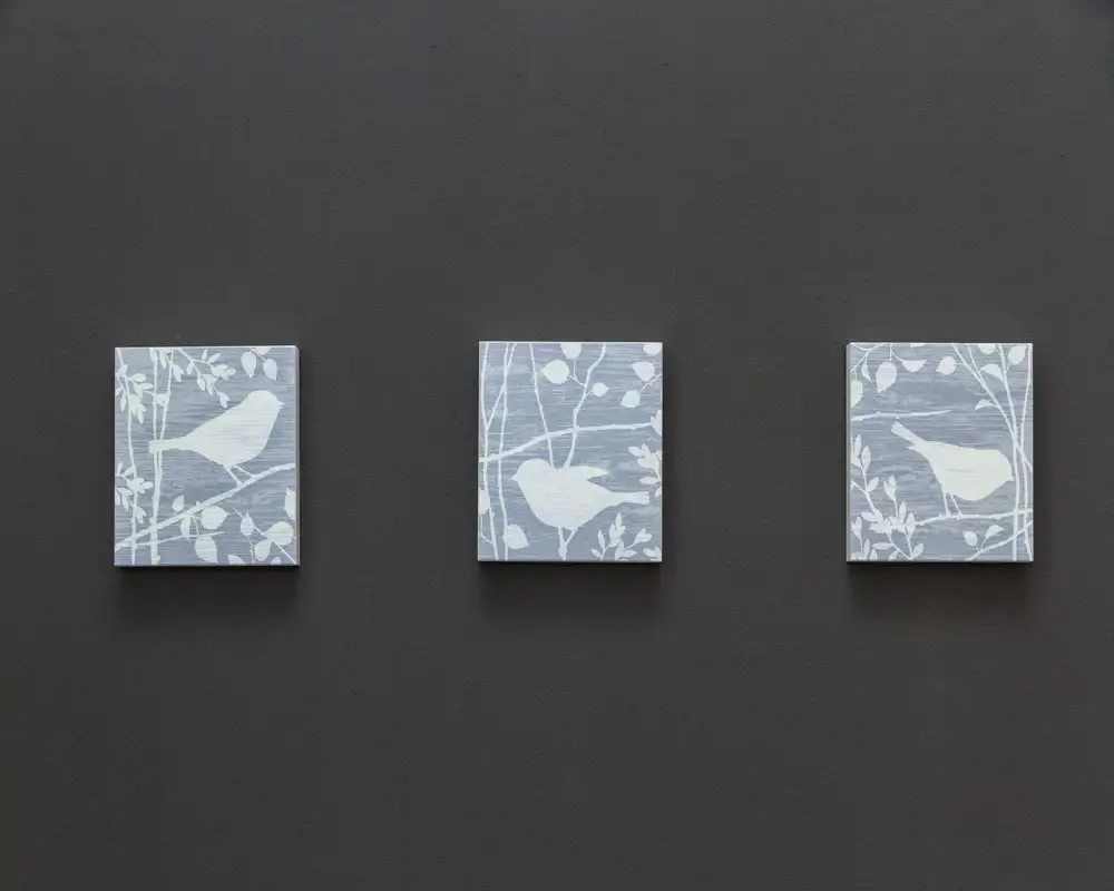 Gros plan de trois petites toiles décoratives représentant des oiseaux accrochées à un mur foncé ; l'image met en avant l'art mural plutôt qu'une pièce identifiable.