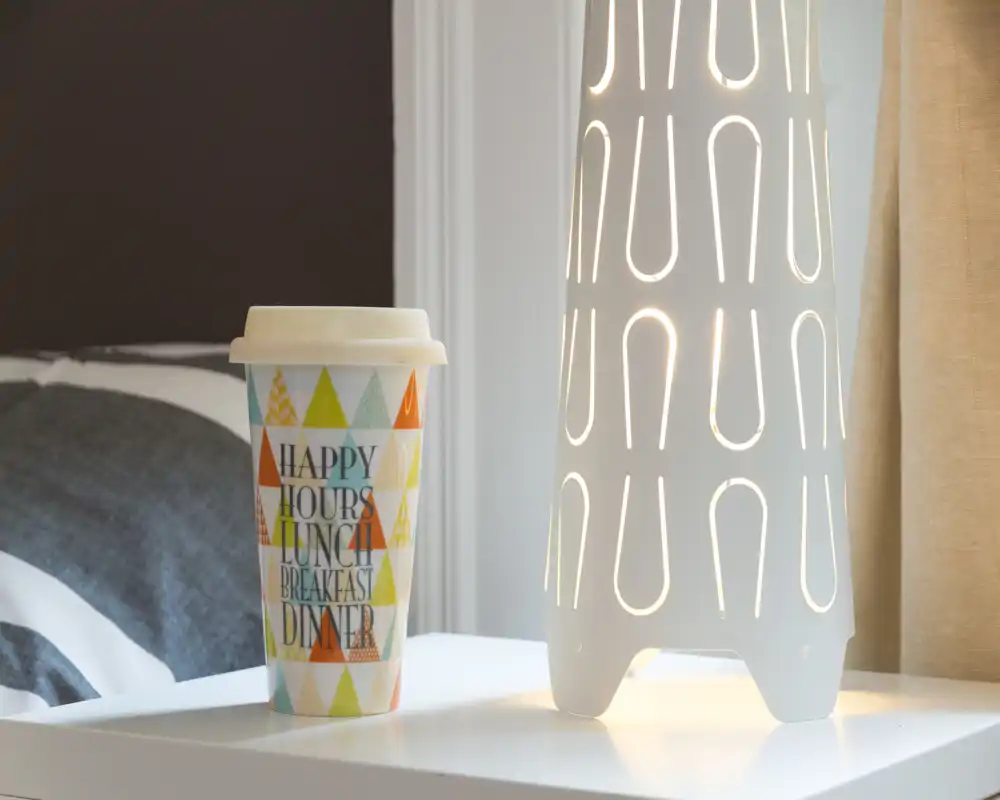 Coin de chambre chaleureux avec une table de chevet, un gobelet décoratif et une lampe moderne perforée diffusant une lumière d'ambiance.