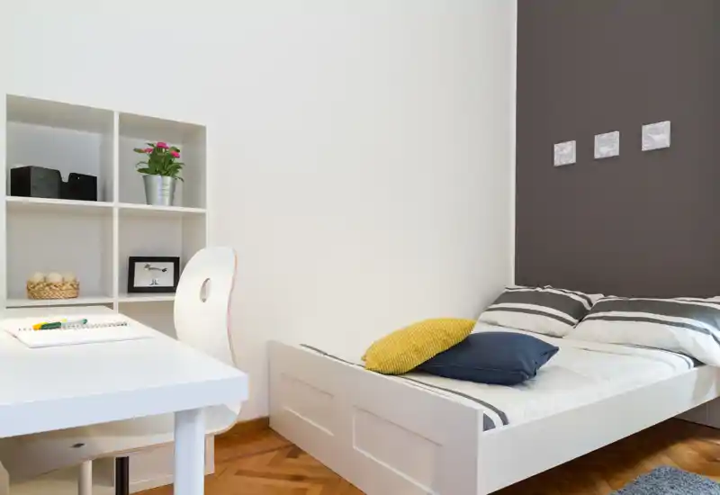 Chambre simple moderne et lumineuse avec lit à cadre blanc, linge de lit rayé et coussins d'accent; comprend un petit bureau blanc et des étagères, idéal pour étudier ou télétravailler.