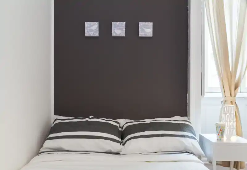 Chambre lumineuse au style épuré avec un lit double bien fait contre un mur foncé, lumière naturelle douce provenant de la fenêtre et table de chevet minimaliste.
