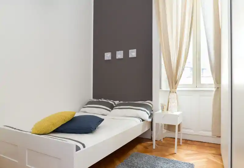Chambre simple lumineuse et soignée avec lit blanc, couette rayée, coussins décoratifs et petite table de chevet près d'une grande fenêtre aux rideaux clairs.