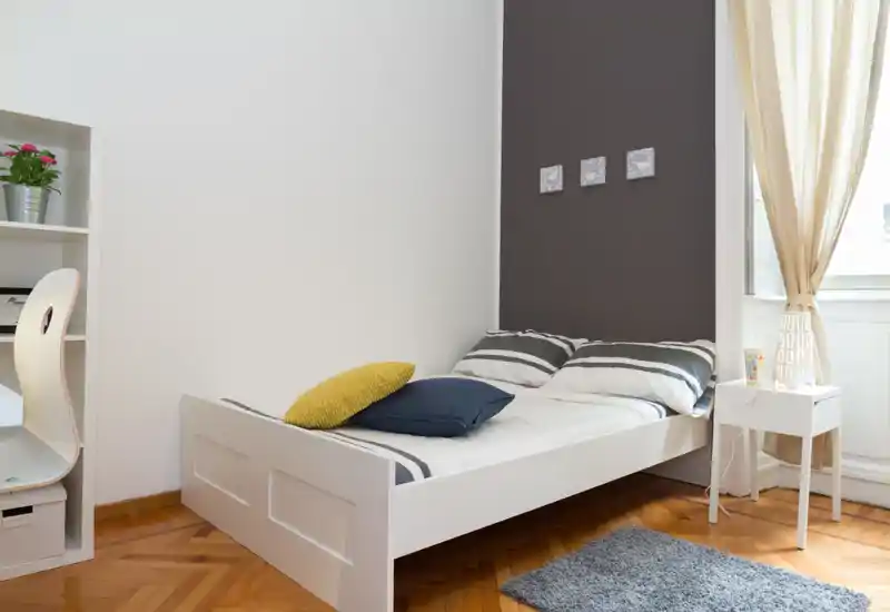 Chambre lumineuse et minimaliste avec un lit blanc une place, des oreillers rayés, une petite table de chevet et la lumière naturelle venant de la fenêtre. Lignes épurées et textiles doux pour une ambiance moderne et chaleureuse.