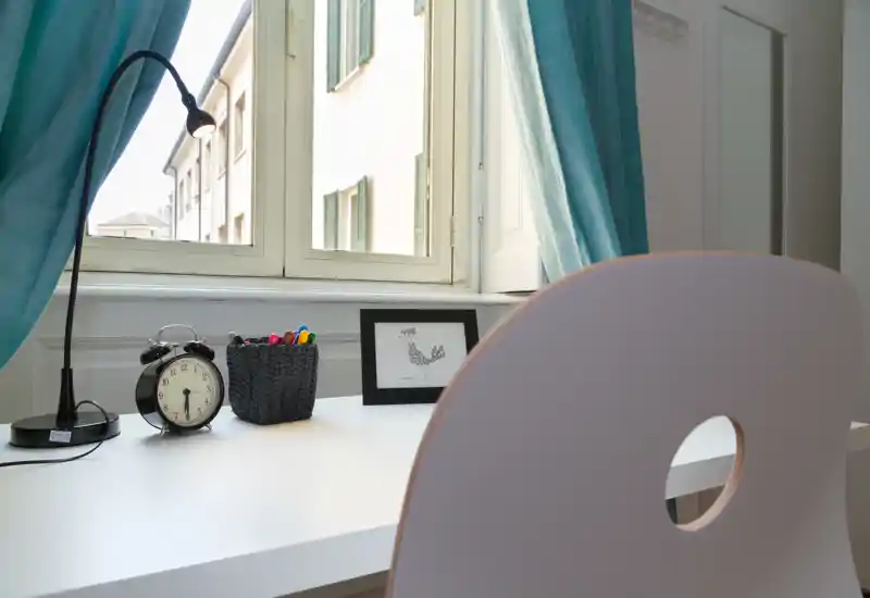 Coin bureau lumineux près d'une fenêtre avec un bureau blanc, chaise, lampe de bureau, réveil et petits objets décoratifs ; parfait pour mettre en valeur un espace de travail cosy.