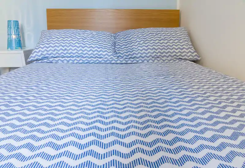 Chambre lumineuse et soignée avec un grand lit recouvert d'une parure bleue et blanche à motifs et une tête de lit en bois simple ; décoration de chevet minimale pour une atmosphère apaisante.