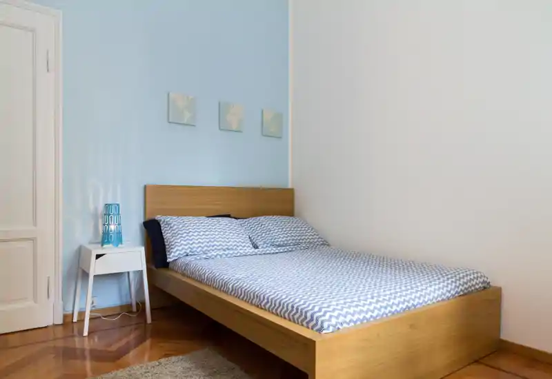 Chambre moderne et lumineuse avec un lit en bois habillé de linge de lit à chevrons bleu, une table de chevet blanche simple et une lampe décorative. Des tableaux muraux pastel et un parquet chaleureux complètent cet espace accueillant.