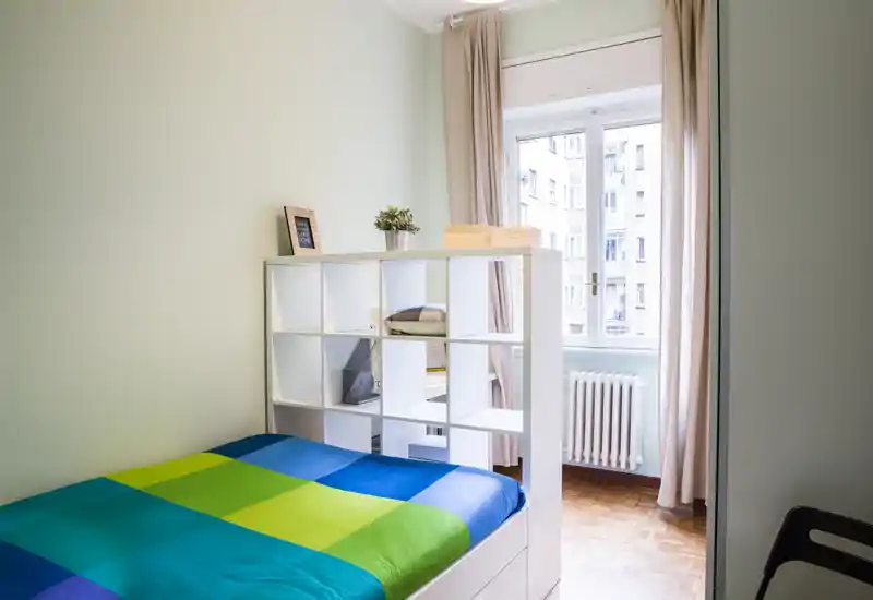 Bright, tidy bedroom with a double bed framed by a white shelving unit that separates the sleeping area from the window. Natural light fills the room and the colorful bedspread gives a fresh, modern feel.