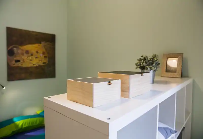 Stylish small bedroom corner with a white shelving unit topped by decorative wooden boxes, a framed print and a small plant; soft bedding is visible at the lower-left corner.