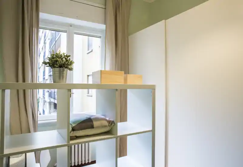 Bright, tidy bedroom corner featuring a freestanding cubed shelf, wardrobe and a window with curtains; decorative plant and cushion add a cozy touch.