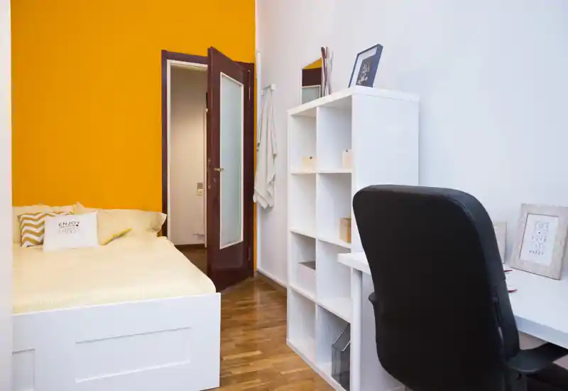 Bright, neatly staged bedroom with a white bed, warm orange accent wall and a compact workspace. Clean wooden floor and shelving make this room ideal for a single occupant or student.