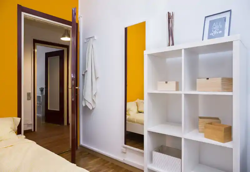 Sunny, tidy bedroom corner with a neatly made bed, full-height mirror and white shelving unit; warm orange accent wall and wooden floor create a cozy, well-lit atmosphere.