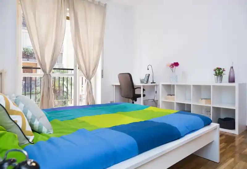 Camera da letto moderna e luminosa con copriletto colorato, scrivania e scaffalature; ampia finestra che garantisce molta luce naturale.