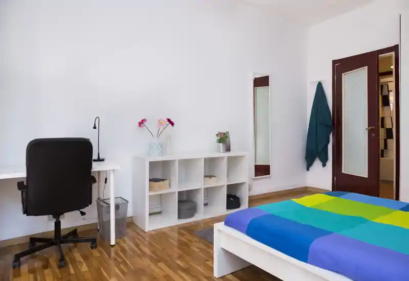 Camera da letto luminosa e ben allestita con letto matrimoniale dai colori vivaci, scrivania e sedia, mensole aperte e dettagli decorativi. Pavimento in legno e arredo minimal conferiscono un aspetto pulito e moderno adatto a annunci di affitto.