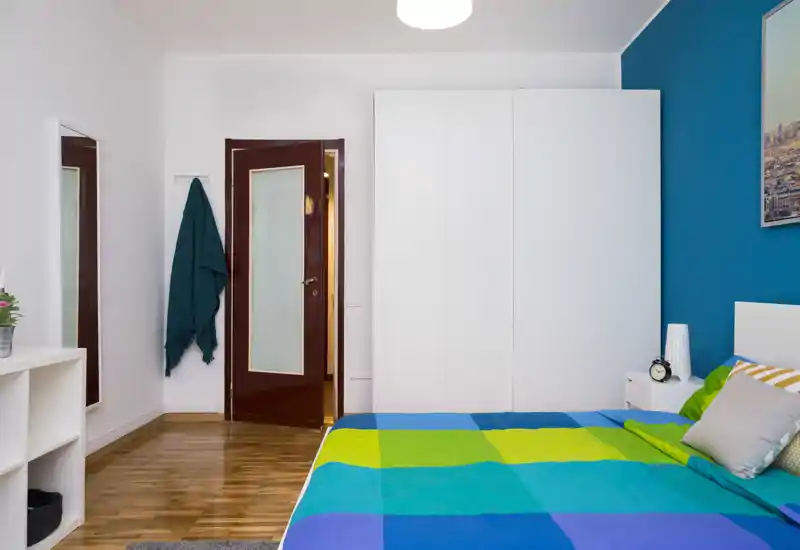 Camera da letto luminosa e ordinata con un letto colorato, armadio bianco e comodino — perfetta per annunci di affitto.