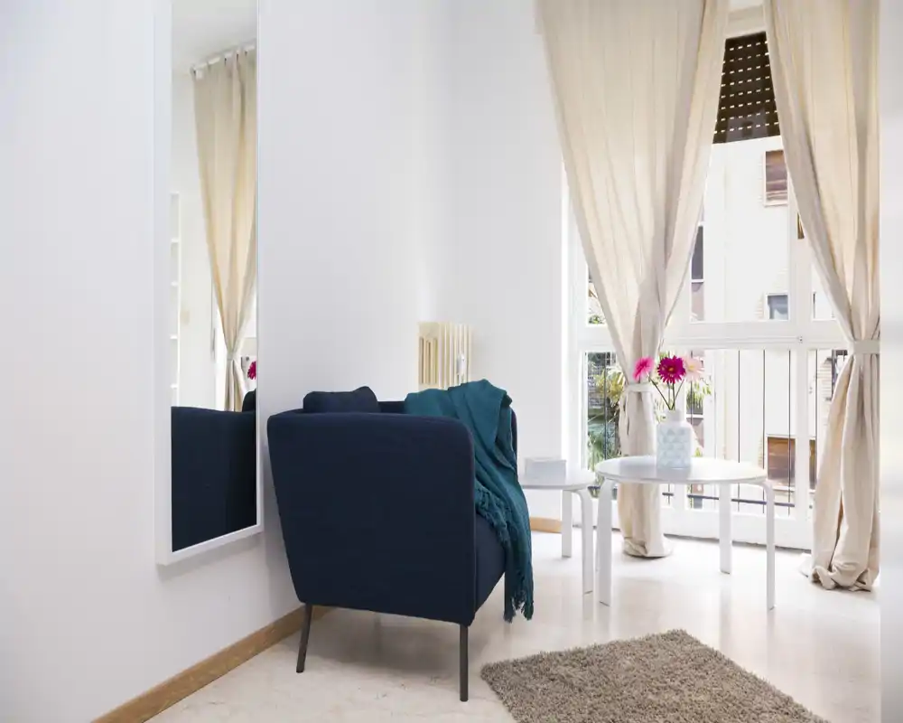 Bright, cozy living area with a single blue armchair, soft rug and a small round table by large windows letting in natural light.